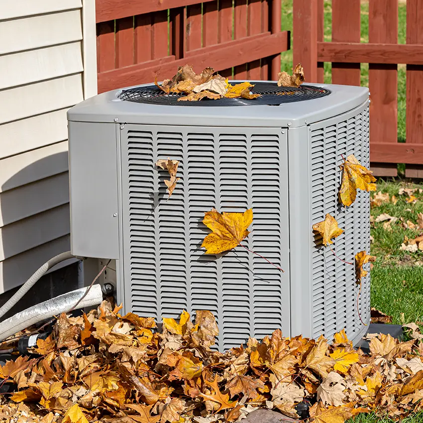 An A/C unit outdoors with Fall leaves around it and stuck in the grates.